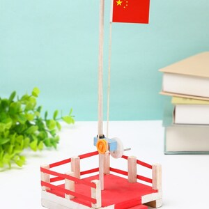 Pode incluir: Uma plataforma de elevação de bandeira de brinquedo de madeira com uma bandeira vermelha com a bandeira chinesa. A plataforma tem 10 cm de comprimento, 11 cm de largura e 28 cm de altura.
