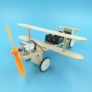 May include: A wooden toy aeroplane with an orange propeller, a small motor, and two wheels. The aeroplane is powered by batteries.