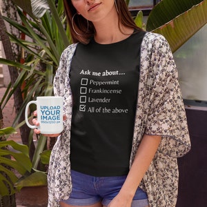 May include: A woman wearing a black t-shirt with the text "Ask me about...." and a checklist of essential oils. She is holding a white mug with the text "UPLOAD YOUR IMAGE" and wearing a floral kimono and denim shorts.