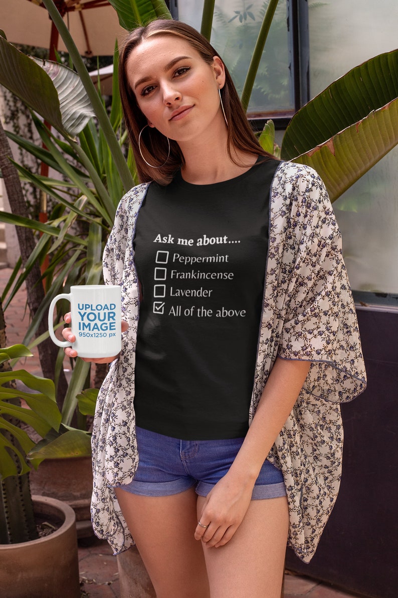 May include: A woman wearing a black t-shirt with the text "Ask me about...." and a checklist of essential oils. She is holding a white mug with the text "UPLOAD YOUR IMAGE" and wearing a floral kimono and denim shorts.
