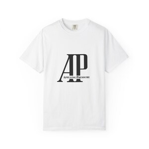 May include: A white short-sleeved t-shirt featuring a large black graphic on the chest. The graphic displays the letters AP in a bold serif font with the words APPLYING PRESSURE printed in a smaller serif font centered below the letters.