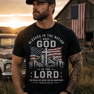 May include: A black short-sleeved t-shirt featuring a graphic print of an American flag, a cross, and mountain silhouettes. The text reads: Blessed is the nation whose God is the Lord, the people he chose for his inheritance, Psalm 33:12.