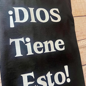 May include: Black t-shirt with the Spanish text "¡DIOS Tiene Esto!" in large, white, bold letters. The text is centered on the shirt. The shirt is folded and appears to be made of a soft material.