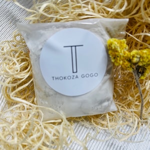Puede incluir: Una pequeña bolsa de plástico transparente llena de un polvo blanco, sellada y etiquetada con una pegatina circular blanca. La pegatina presenta una 'T' negra y las palabras 'THOKOZA GOGO'. La bolsa está metida en paja decorativa con una flor amarilla seca.
