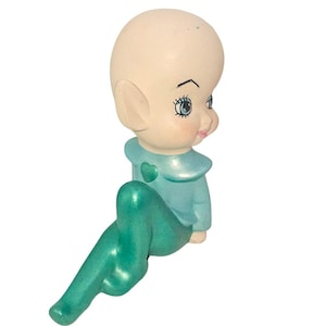 Op de afbeelding: Een keramieken beeldje van een zittende elf met een groot hoofd en puntige oren. De elf draagt een lichtblauwe outfit met een hartdetail en turquoise leggings. Het beeldje heeft een bleke teint en gedetailleerde gelaatstrekken.