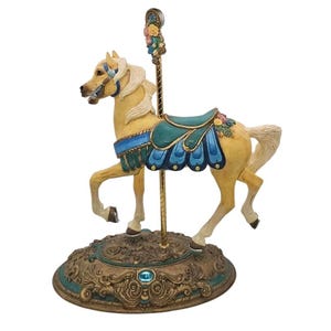 May include: A carousel horse figurine in a trotting pose. The horse is painted in a cream color with a flowing mane and tail. It has a blue saddle and bridle, and is mounted on a decorative base with floral accents.