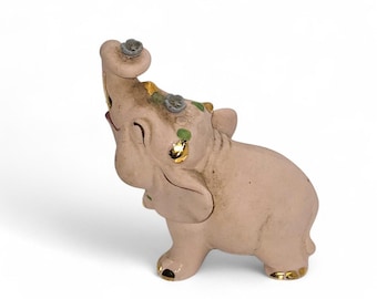 May include: A small, pink ceramic elephant figurine with its trunk raised. The elephant has gold accents on its ears, feet, and trunk. It is decorated with small green and gray floral details. The figurine is set against a white background.