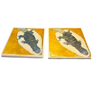May include: Two square decorative tiles. Each tile features a lizard illustration in blue and gold tones. The lizard is centered on a cream-colored shape, set against a gold background. The tiles have a wooden frame.