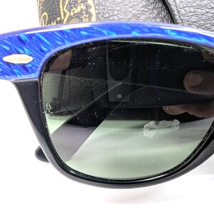 Op de afbeelding: Zwarte zonnebril met een blauw gemarmerd bovenframe en groene glazen. De zonnebril heeft een klassiek ontwerp met een rechthoekige vorm. De tekst "RAY-BAN U.S.A." is zichtbaar op de lens.