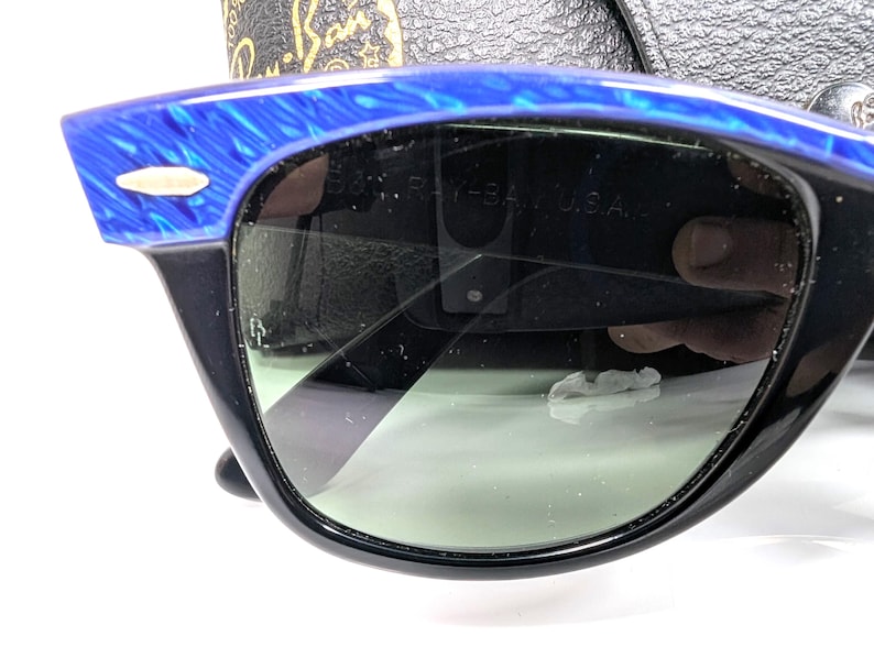 Op de afbeelding: Zwarte zonnebril met een blauw gemarmerd bovenframe en groene glazen. De zonnebril heeft een klassiek ontwerp met een rechthoekige vorm. De tekst "RAY-BAN U.S.A." is zichtbaar op de lens.