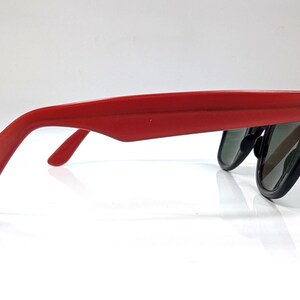 May include: Red sunglasses with black frames and dark lenses. The sunglasses are viewed from the side, showcasing the red arms and the shape of the lenses. The design is a classic style, suitable for sun protection.