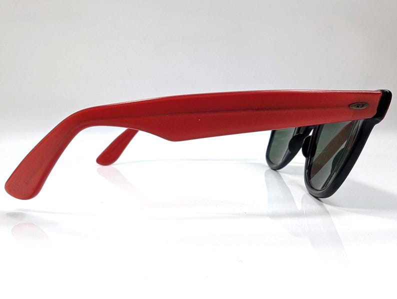 May include: Red sunglasses with black frames and dark lenses. The sunglasses are viewed from the side, showcasing the red arms and the shape of the lenses. The design is a classic style, suitable for sun protection.