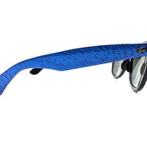 Op de afbeelding: Ray-Ban zonnebril met blauw patroon op de pootjes en zwarte montuur. Het Ray-Ban logo is zichtbaar aan de zijkant. De glazen zijn groen en reflecterend.