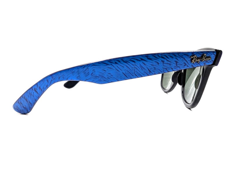 Op de afbeelding: Ray-Ban zonnebril met blauw patroon op de pootjes en zwarte montuur. Het Ray-Ban logo is zichtbaar aan de zijkant. De glazen zijn groen en reflecterend.