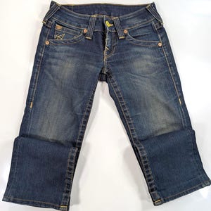 Pode incluir: Jeans de ganga azul escuro com um design cl&aacute;ssico de cinco bolsos. Apresenta fecho de bot&atilde;o e z&iacute;per, presilhas para cinto e costuras decorativas. Os jeans s&atilde;o dobrados na parte inferior, mostrando a textura e as varia&ccedil;&otilde;es de cor da ganga.
