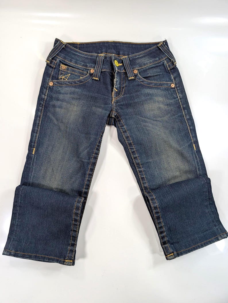 Pode incluir: Jeans de ganga azul escuro com um design cl&aacute;ssico de cinco bolsos. Apresenta fecho de bot&atilde;o e z&iacute;per, presilhas para cinto e costuras decorativas. Os jeans s&atilde;o dobrados na parte inferior, mostrando a textura e as varia&ccedil;&otilde;es de cor da ganga.