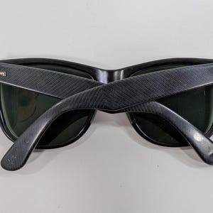 Op de afbeelding: Zwarte Ray-Ban zonnebril met een textuur van diagonale lijnen op de pootjes. Het montuur heeft donkergroene glazen en het Ray-Ban logo is in goudkleurig metaal bevestigd op het buitenste scharnier van elk pootje.