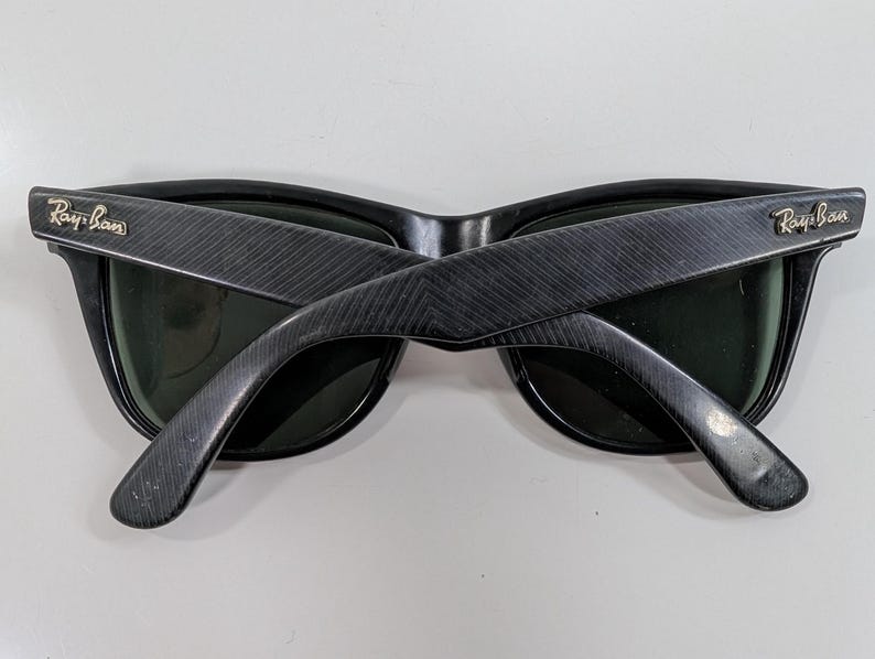 Op de afbeelding: Zwarte Ray-Ban zonnebril met een textuur van diagonale lijnen op de pootjes. Het montuur heeft donkergroene glazen en het Ray-Ban logo is in goudkleurig metaal bevestigd op het buitenste scharnier van elk pootje.