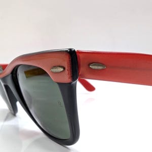 May include: Close-up of retro-style sunglasses with a black frame and red arms. The lenses are a dark green color. The sunglasses have a classic, vintage design, with a slight cat-eye shape.