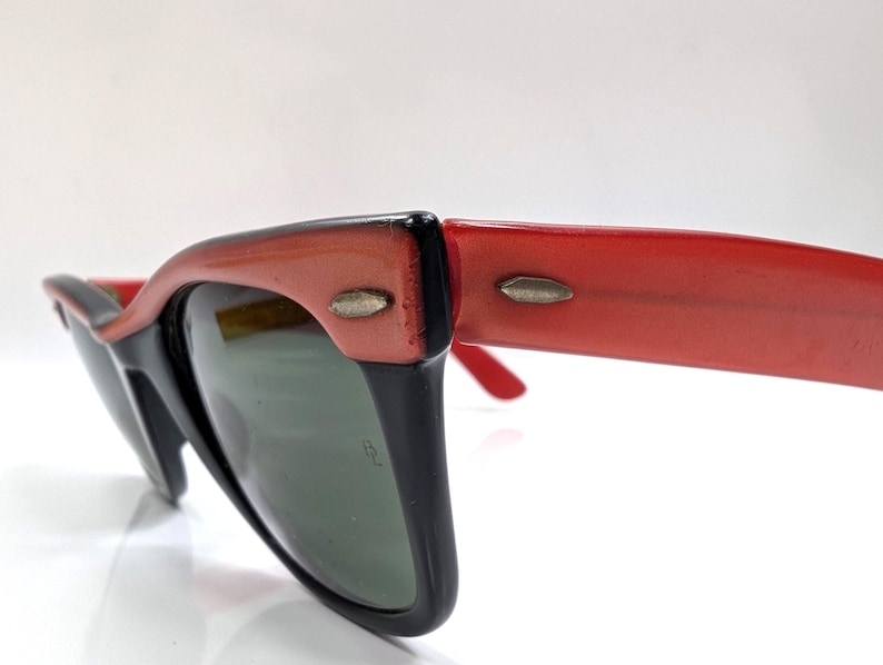 May include: Close-up of retro-style sunglasses with a black frame and red arms. The lenses are a dark green color. The sunglasses have a classic, vintage design, with a slight cat-eye shape.