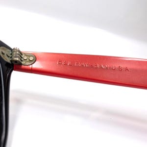 May include: Close-up of vintage Ray-Ban sunglasses. The black frame contrasts with the red arm, which features the text "B&L RAY-BAN U.S.A." The hinge is a gold color. The lenses are a dark green.