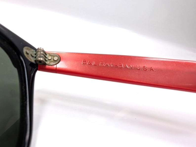 May include: Close-up of vintage Ray-Ban sunglasses. The black frame contrasts with the red arm, which features the text "B&L RAY-BAN U.S.A." The hinge is a gold color. The lenses are a dark green.