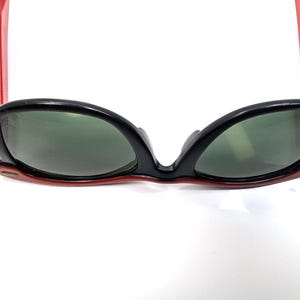 May include: Black sunglasses with red arms and green lenses. The sunglasses have a retro cat-eye shape. The frame is black with red accents. The lenses are a dark green color.