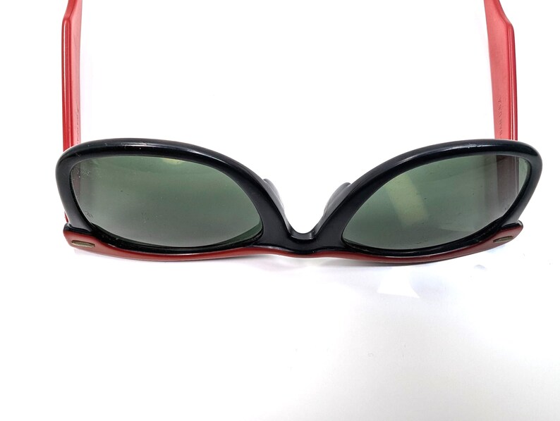 May include: Black sunglasses with red arms and green lenses. The sunglasses have a retro cat-eye shape. The frame is black with red accents. The lenses are a dark green color.