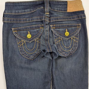 Pode incluir: Jeans de ganga azul escuro com costuras amarelas e bolsos traseiros decorativos. Os jeans apresentam um remendo de couro e fechos de bot&atilde;o amarelos. Os bolsos traseiros t&ecirc;m um design &uacute;nico.