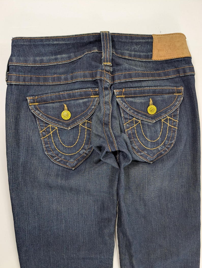 Pode incluir: Jeans de ganga azul escuro com costuras amarelas e bolsos traseiros decorativos. Os jeans apresentam um remendo de couro e fechos de bot&atilde;o amarelos. Os bolsos traseiros t&ecirc;m um design &uacute;nico.