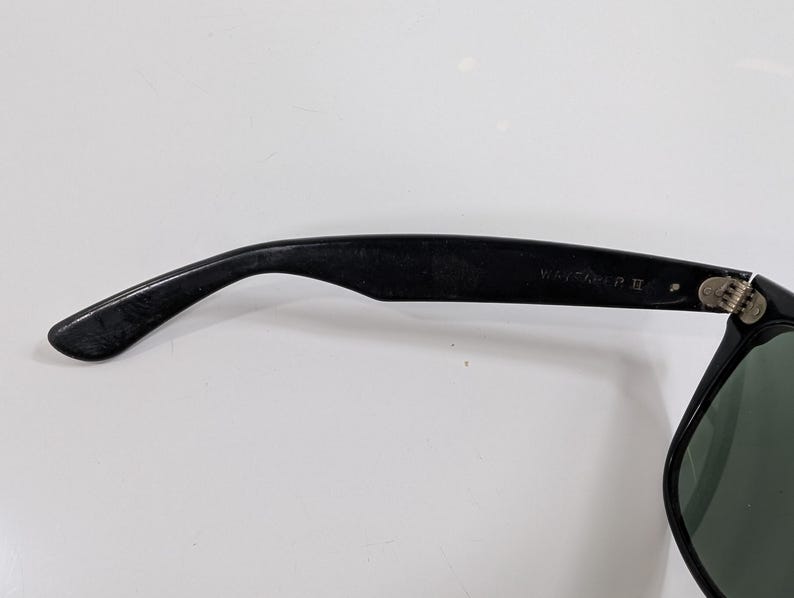 Op de afbeelding: Zwarte zonnebril met een gebogen brilpoot, voorzien van een metalen scharnier en de tekst WAYFARER II in reli&euml;f aan de binnenzijde. Het montuur is glanzend zwart met een donkergroene lens zichtbaar aan de rechterkant van de bril.