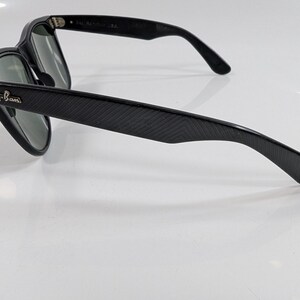 Op de afbeelding: Zwarte zonnebril met een textuur van diagonale lijnen op de pootjes. De monturen hebben een zilverkleurig Ray-Ban logo op het buitenste scharnier. De glazen zijn donkergroen en de binnenkant van de pootjes toont de tekst Ray-Ban USA.