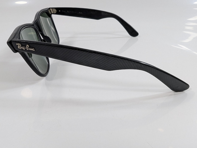 Op de afbeelding: Zwarte zonnebril met een textuur van diagonale lijnen op de pootjes. De monturen hebben een zilverkleurig Ray-Ban logo op het buitenste scharnier. De glazen zijn donkergroen en de binnenkant van de pootjes toont de tekst Ray-Ban USA.
