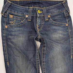 Pode incluir: Close-up de um par de jeans de ganga de lavagem escura. Os jeans apresentam um fecho de bot&atilde;o, bolsos frontais e traseiros e costuras decorativas amarelas contrastantes. Os jeans t&ecirc;m um aspeto cl&aacute;ssico e usado.