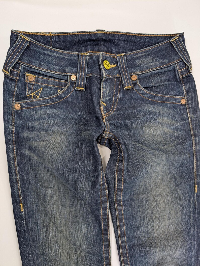 Pode incluir: Close-up de um par de jeans de ganga de lavagem escura. Os jeans apresentam um fecho de bot&atilde;o, bolsos frontais e traseiros e costuras decorativas amarelas contrastantes. Os jeans t&ecirc;m um aspeto cl&aacute;ssico e usado.