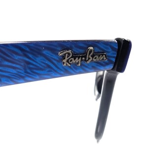 Op de afbeelding: Close-up van een Ray-Ban zonnebril. Het montuur is helderblauw met een gemarmerd patroon en het Ray-Ban logo is in zilver weergegeven. De pootjes zijn zwart. De zonnebril staat tegen een witte achtergrond.