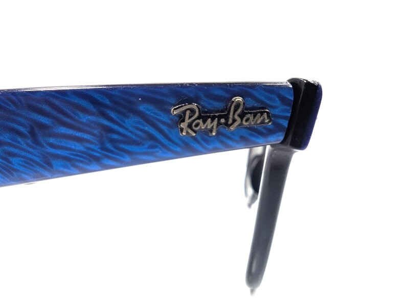 Op de afbeelding: Close-up van een Ray-Ban zonnebril. Het montuur is helderblauw met een gemarmerd patroon en het Ray-Ban logo is in zilver weergegeven. De pootjes zijn zwart. De zonnebril staat tegen een witte achtergrond.