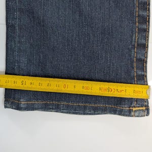 Pode incluir: Close-up de jeans azul escuro com uma fita m&eacute;trica amarela colocada na bainha. A fita mostra medidas em cent&iacute;metros, destacando a largura da abertura da perna da cal&ccedil;a jeans. Os jeans t&ecirc;m costuras vis&iacute;veis.