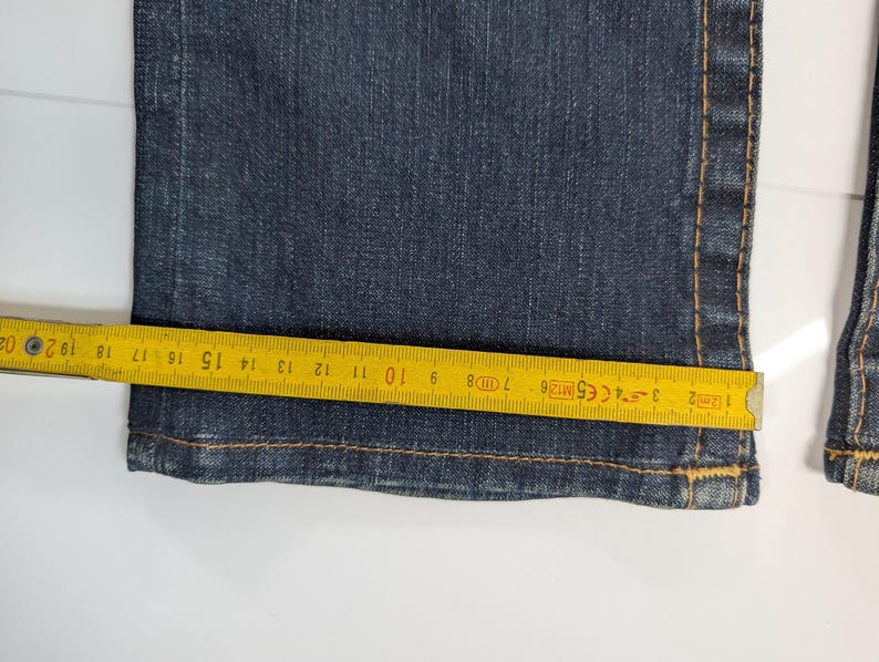 Pode incluir: Close-up de jeans azul escuro com uma fita m&eacute;trica amarela colocada na bainha. A fita mostra medidas em cent&iacute;metros, destacando a largura da abertura da perna da cal&ccedil;a jeans. Os jeans t&ecirc;m costuras vis&iacute;veis.