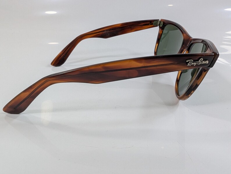 Op de afbeelding: Zonnebril met schildpadpatroon en een bruin en amberkleurig gemarmerd montuur. De pootjes zijn bij het scharnier voorzien van een zilverkleurig Ray-Ban logo. De glazen zijn donkergroen en bieden zicht op de interne structuur van het montuur.