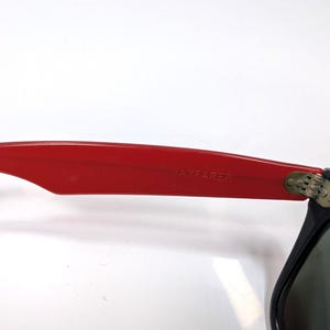 May include: Close-up of black sunglasses with green lenses. The red arm of the sunglasses has the text "AYFARER" printed on it. The sunglasses are set against a white background.