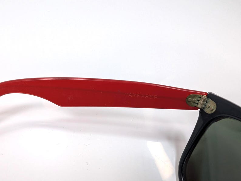 May include: Close-up of black sunglasses with green lenses. The red arm of the sunglasses has the text "AYFARER" printed on it. The sunglasses are set against a white background.