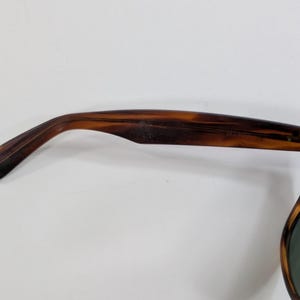 Op de afbeelding: Zonnebril met schildpadpatroon met een gebogen pootje met een zichtbaar metalen scharnier en schroef. De binnenkant van het pootje toont de in reli&euml;f aangebrachte tekst WAYFARER, terwijl de donkergroene lens aan de rechterkant zichtbaar is.