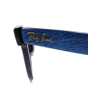 Op de afbeelding: Close-up van een Ray-Ban zonnebril. De monturen zijn zwart met een blauw gemarmerd patroon op de pootjes. Het Ray-Ban logo is zichtbaar op de poot van de bril. De bril staat tegen een witte achtergrond.