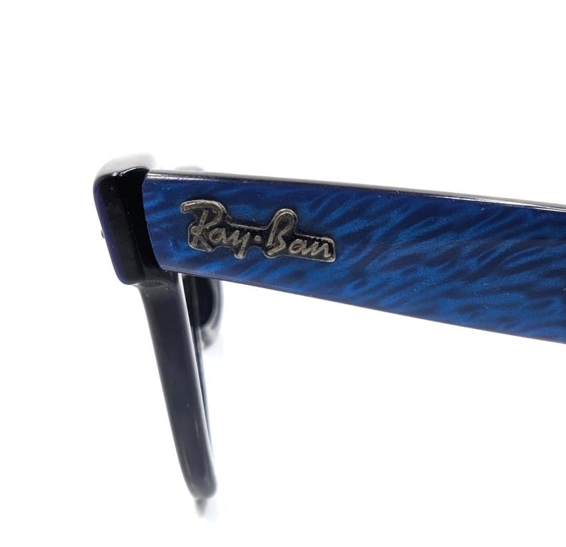 Op de afbeelding: Close-up van een Ray-Ban zonnebril. De monturen zijn zwart met een blauw gemarmerd patroon op de pootjes. Het Ray-Ban logo is zichtbaar op de poot van de bril. De bril staat tegen een witte achtergrond.