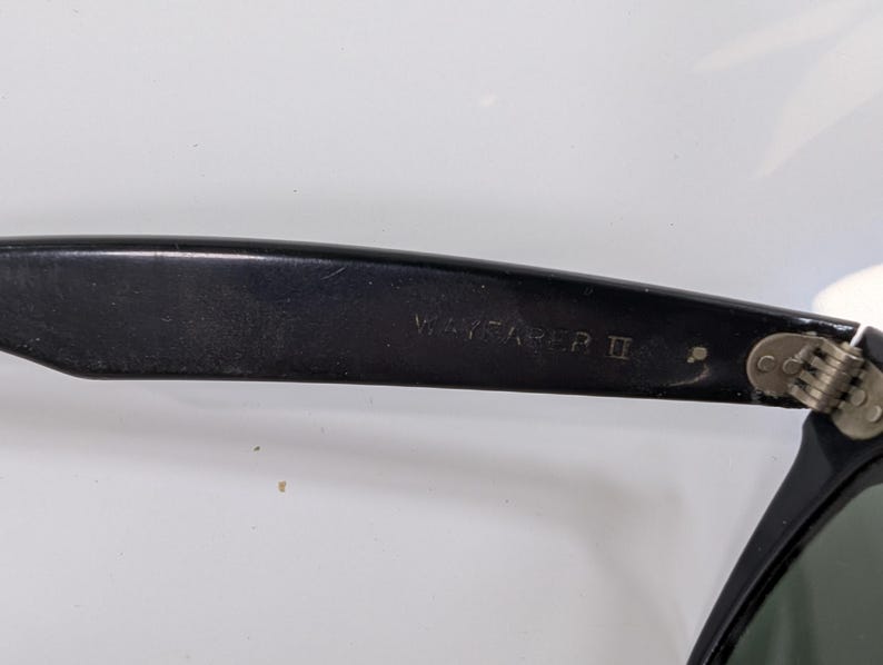 Op de afbeelding: Zwarte kunststof zonnebrilpoot met de tekst WAYFARER II in reli&euml;f aan de binnenzijde. De poot is met het montuur verbonden door een metalen scharnier met zichtbare schroeven, en een donkergroene lens is rechtsonder gedeeltelijk zichtbaar.