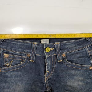 Pode incluir: Close-up de um par de jeans de ganga azul escura. Os jeans apresentam um fecho de bot&atilde;o, bolsos frontais e traseiros e pespontos amarelos. Uma fita m&eacute;trica amarela &eacute; colocada na parte superior dos jeans, mostrando uma medida de aproximadamente 43 cm.