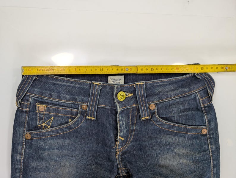 Pode incluir: Close-up de um par de jeans de ganga azul escura. Os jeans apresentam um fecho de bot&atilde;o, bolsos frontais e traseiros e pespontos amarelos. Uma fita m&eacute;trica amarela &eacute; colocada na parte superior dos jeans, mostrando uma medida de aproximadamente 43 cm.