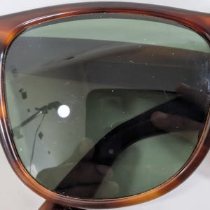 Op de afbeelding: Zonnebril met schildpadpatroon en een afgerond montuur met een bruine en zwarte gemarmerde afwerking. De glazen zijn donkergroen getint, met een klein zilverkleurig metalen accent zichtbaar op de bovenhoek van het montuur bij het scharnier.