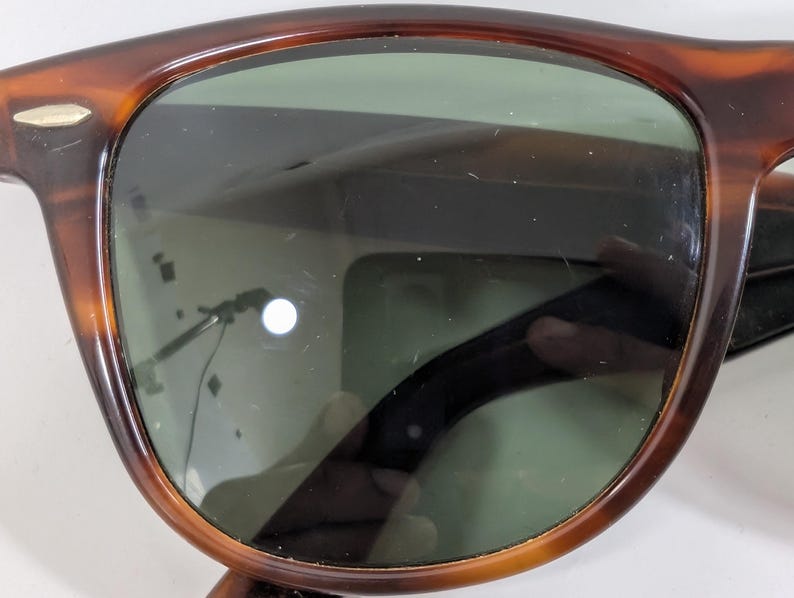 Op de afbeelding: Zonnebril met schildpadpatroon en een afgerond montuur met een bruine en zwarte gemarmerde afwerking. De glazen zijn donkergroen getint, met een klein zilverkleurig metalen accent zichtbaar op de bovenhoek van het montuur bij het scharnier.